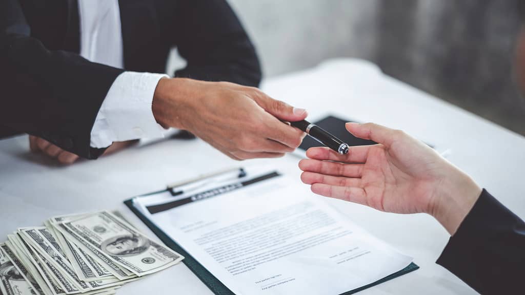 person handing a pen to sign the contract with money on the table